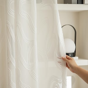 Puede incluir: Cortina blanca transparente con un patrón ondulado bordado. La cortina se mantiene abierta, revelando un vistazo de una estantería con libros y una lámpara. La tela es ligera y permite que la luz se filtre.