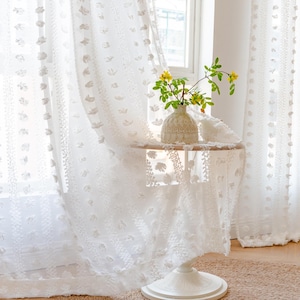 May include: White sheer curtains with a textured, floral pattern and pom-poms. The curtains are hanging in a window and a vase of yellow flowers is on a white table in front of them.