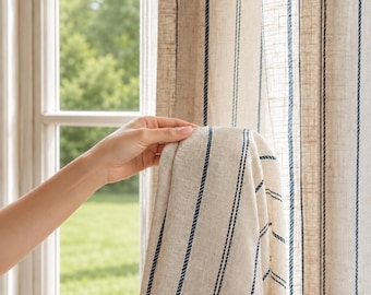 May include: A beige curtain panel featuring vertical navy blue pinstripes. The fabric has a visible woven texture and hangs in soft folds, allowing light to pass through the material. The design consists of repeating thin blue lines.