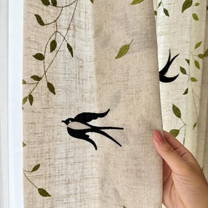 May include: A beige curtain panel featuring an embroidered pattern of green leafy vines and black swallow birds. The fabric has a visible woven texture, and the embroidery is distributed across the surface, creating a nature-inspired design.