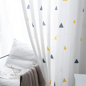 Puede incluir: Cortinas blancas y transparentes con un patrón repetitivo de triángulos amarillos y azul marino. Las cortinas cuelgan frente a una ventana. Una silla y una manta son visibles en primer plano. Estas cortinas añaden un toque moderno a cualquier habitación.
