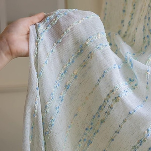 May include: Sheer white fabric with a delicate texture, adorned with rows of small, colorful embellishments in blue, yellow, and green. The fabric is held up, showcasing its lightweight and airy quality, ideal for curtains or decorative purposes.