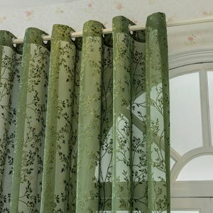 Könnte beinhalten: Grüne transparente Vorhänge mit einem zarten Baumzweigmuster. Die Vorhänge hängen an einer weißen Stange und lassen Licht durchscheinen. Das Design zeigt ein sich wiederholendes Muster aus Blättern und Zweigen.
