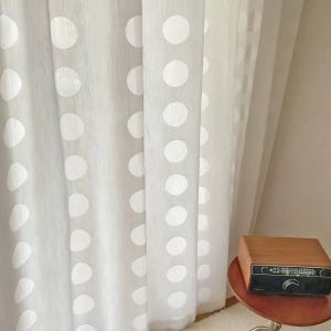 Op de afbeelding: Transparante witte gordijnen met een patroon van grote witte stippen. Het licht schijnt door de gordijnen en creëert een zacht, diffuus effect. Een vintage houten radio staat op een kleine, ronde tafel.