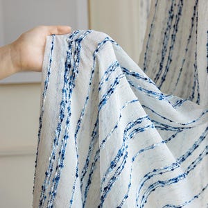 May include: White fabric with vertical blue lines and dotted accents. The fabric is held up, showing its texture and pattern. The blue lines vary in thickness and spacing, creating a textured effect. The fabric appears to be lightweight and suitable for curtains or other home decor.