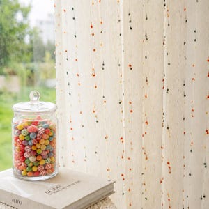 May include: A clear glass jar filled with multicolored round candies sits on a beige book titled at 100. Beside it hangs a cream-colored curtain featuring vertical rows of small, textured tufts in shades of orange, dark green, and black.