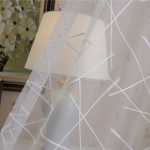 May include: Sheer white curtain with a geometric pattern of intersecting lines. The curtain is partially covering a lamp with a white lampshade. In the background, a framed floral painting and books are visible.