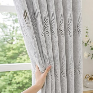 Puede incluir: Un panel de cortina gris claro con un patrón vertical repetitivo de formas texturizadas y elevadas similares a hojas. La tela tiene un acabado mate suave con pliegues profundos, mostrando un diseño consistente en toda la superficie.