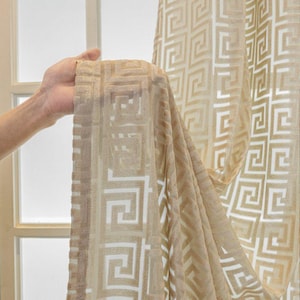 以下が含まれることがあります： 幾何学的なギリシャキー模様のベージュのシアーカーテン。カーテンは軽量の生地で作られており、光を通します。デザインは、より明るいベージュの色合いの繰り返しの正方形パターンです。
