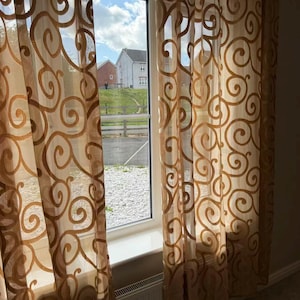 May include: Sheer window curtains featuring a repeating golden-brown swirl pattern on a translucent fabric. The curtains hang vertically on either side of a window, allowing natural light to filter through the semi-transparent material.