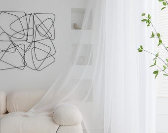 May include: White sheer curtains hanging in a room with a white couch and a black geometric wall art piece.