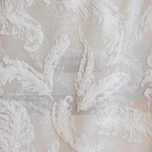 Puede incluir: Primer plano de una tela blanca con un patrón texturizado en relieve de plumas estilizadas. Las plumas densas crean un efecto tridimensional sobre el fondo transparente. La tela tiene una calidad delicada y aireada.