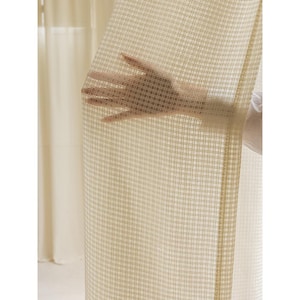 May include: A sheer, beige curtain with a grid pattern is held by a hand. The curtain is semi-transparent, allowing light to pass through. The grid pattern is made up of thin, light brown lines. The background shows a second, solid beige curtain.