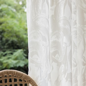 May include: White curtains featuring a large, abstract brushstroke design. The curtains are in front of a window with a view of green foliage. A woven chair is in the foreground.