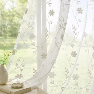 May include: Sheer white curtain panels with embroidered floral and leaf designs. The delicate embroidery features a light beige color, creating a subtle contrast against the white fabric. The curtains are hanging in front of a window, with a blurred green background.