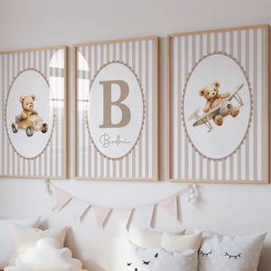 May include: Three framed nursery art prints. Each print features a beige teddy bear illustration. One print shows a teddy bear in a car, another with a plane, and the third with a large letter "B" and the name "Bodhi". The background is striped.