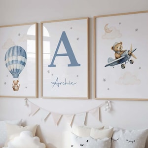 May include: Three framed art prints with a nursery theme. One print features a blue hot air balloon with a teddy bear, another has a large blue letter "A" with the name "Archie", and the third shows a teddy bear in a biplane. The prints have a light wood frame.