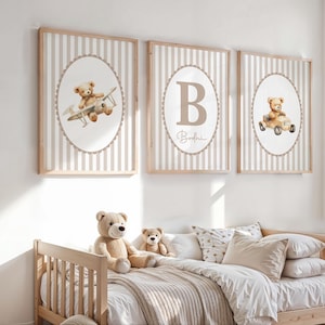 May include: Three framed nursery art prints with a teddy bear theme. Each print features a beige and white striped background with an oval illustration of a teddy bear in a plane, car, and the letter "B". The prints are in wooden frames.
