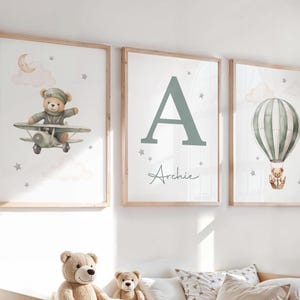 May include: Three framed art prints for a nursery. One depicts a teddy bear in a biplane, another has a large letter "A" with the name "Archie", and the third shows a hot air balloon with a teddy bear. The prints have a soft colour palette.