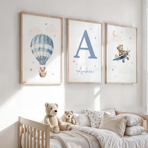 May include: Three framed nursery art prints. One features a blue and white hot air balloon with a teddy bear. Another displays a large blue letter "A" with the name "Archie". The third shows a teddy bear piloting a blue airplane.