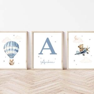 May include: Three framed nursery art prints. One features a blue and white hot air balloon with a teddy bear. Another displays the letter "A" and the name "Archie". The third shows a teddy bear in a plane.