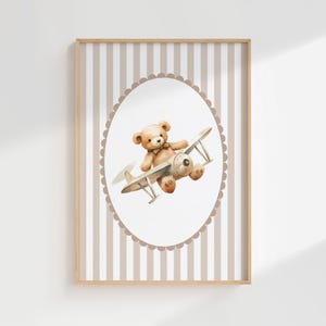 May include: A framed print with a watercolour illustration of a teddy bear flying a vintage biplane. The bear is light brown, and the plane is beige and grey. The background has vertical stripes in beige and white, with a scalloped oval frame.
