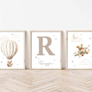 Pode incluir: Tr&ecirc;s impress&otilde;es art&iacute;sticas emolduradas para o quarto infantil. A primeira apresenta um bal&atilde;o de ar quente com um ursinho de pel&uacute;cia, a segunda uma grande letra "R" com o nome "Reggie", e a terceira um ursinho de pel&uacute;cia num biplano. Todas as impress&otilde;es t&ecirc;m uma paleta de cores neutra.