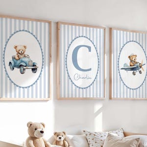 May include: Three framed prints for a nursery, each with a light blue and white striped background. One print shows a teddy bear in a blue car, another has the letter "C" and the name "Charlie", and the third has a teddy bear in a blue aeroplane.