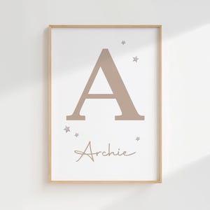 Pode incluir: Impress&atilde;o emoldurada com uma grande letra "A" em tom taupe e o nome "Archie" em escrita cursiva. Pequenas estrelas adornam a letra. A impress&atilde;o tem um fundo branco e uma moldura de madeira clara, ideal para um quarto infantil.
