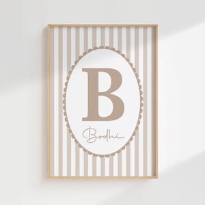 May include: A framed print with a large, light brown letter "B" inside an oval frame with a scalloped edge. The name "Bodhi" is written below. The background features vertical beige and white stripes, all within a wooden frame.