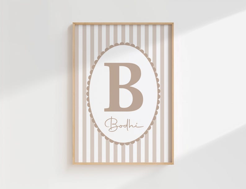 May include: A framed print with a large, light brown letter "B" inside an oval frame with a scalloped edge. The name "Bodhi" is written below. The background features vertical beige and white stripes, all within a wooden frame.