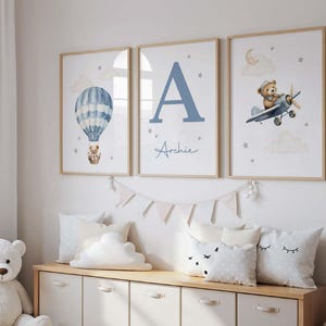 May include: Three framed nursery art prints. One features a blue hot air balloon with a teddy bear, another a large blue letter "A" with the name "Archie", and the third a teddy bear in a plane. The prints have a light wood frame.