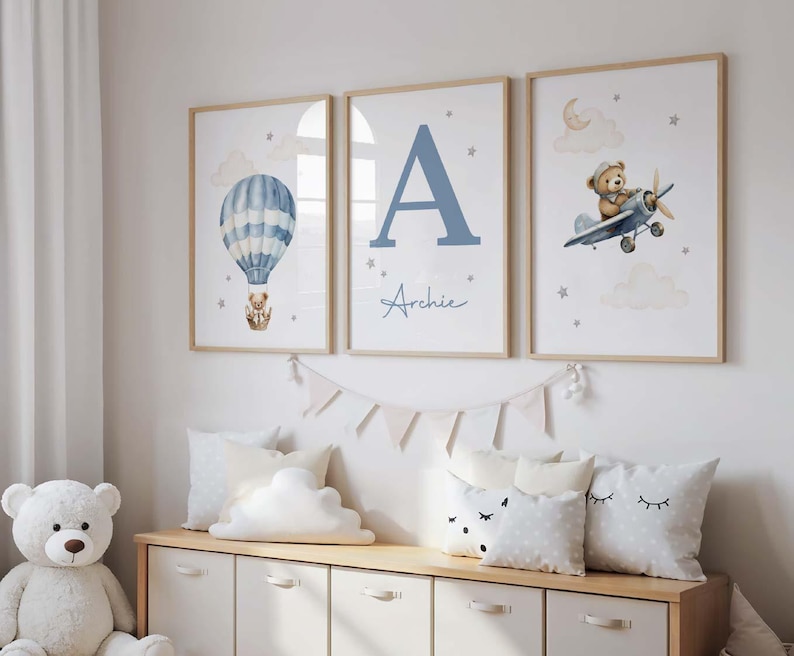 May include: Three framed nursery art prints. One features a blue hot air balloon with a teddy bear, another a large blue letter "A" with the name "Archie", and the third a teddy bear in a plane. The prints have a light wood frame.