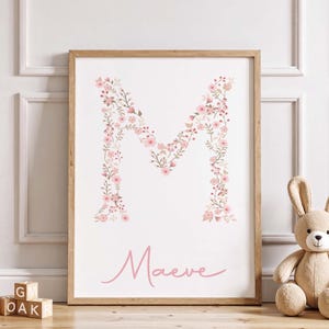 Könnte beinhalten: Gerahmter Druck mit dem Buchstaben "M", gebildet aus rosa Blumen. Der Name "Maeve" steht darunter in rosa Schrift. Ein Stoffhase und Holzklötze ergänzen die Szene, passend für ein Kinderzimmer.