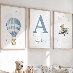 May include: Three framed nursery art prints. One features a blue hot air balloon with a teddy bear, another a large blue letter "A" with the name "Archie", and the third a teddy bear in a plane. The prints have a light wood frame.
