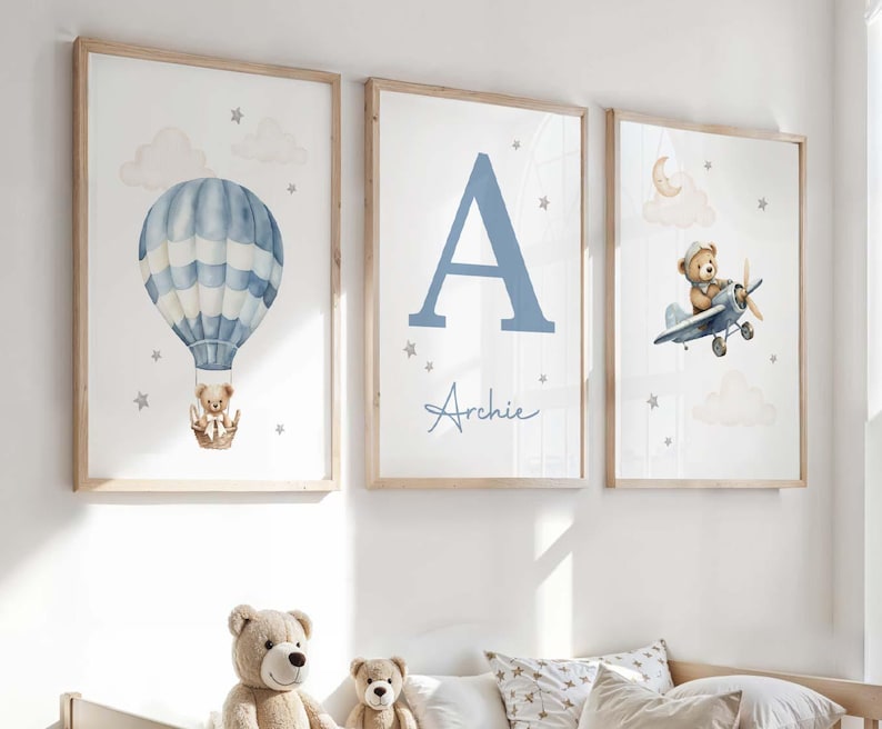 May include: Three framed nursery art prints. One features a blue hot air balloon with a teddy bear, another a large blue letter "A" with the name "Archie", and the third a teddy bear in a plane. The prints have a light wood frame.