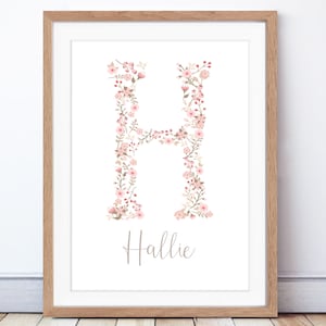 May include: A framed floral print with the letter "H" formed from pink and cream flowers and foliage. The name "Hallie" is written below in cursive. The print is presented in a wooden frame, suitable for a nursery or bedroom.