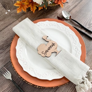 Puede incluir: Una tarjeta de lugar de madera en forma de pavo con el nombre "Savannah" grabado en ella. La tarjeta de lugar está sobre un plato blanco con un plato de carga marrón debajo. Una servilleta blanca está doblada junto a la tarjeta de lugar.