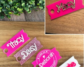 May include: Five pink and white personalised name tags with gold lettering. The tags are shaped like banners and have a hole at the top for hanging. The names on the tags are Tracy, Daisy, Deborah, Cate, and Macy.