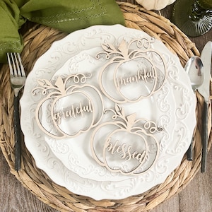 Peut inclure: Trois marque-places en forme de citrouille en bois avec les mots "grateful", "thankful" et "blessed" inscrits dessus. Les marque-places sont sur une assiette blanche avec un dessous de plat en osier.