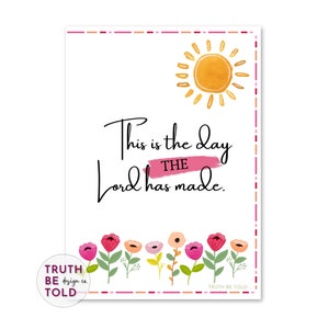 May include: A white print with the text "This is the day the Lord has made." The print features a watercolour sun, a pink banner, and colourful flowers at the bottom. The words "TRUTH BE design co. TOLD" are also printed on the image.