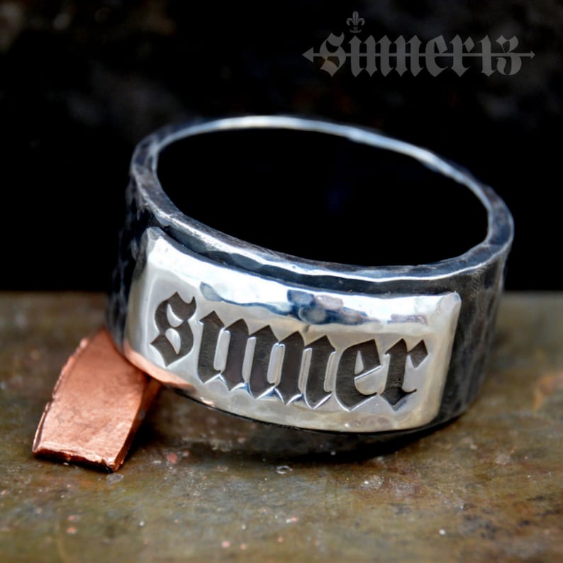 May include: A silver ring with a hammered finish and a rectangular plaque engraved with the word "Summer" in a gothic font.