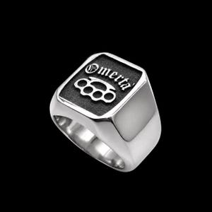 May include: A silver-toned signet ring featuring a rectangular face with a textured black inlay. The face displays the word Omertà in a gothic-style font positioned above a raised graphic of brass knuckles, all rendered in a polished silver finish.