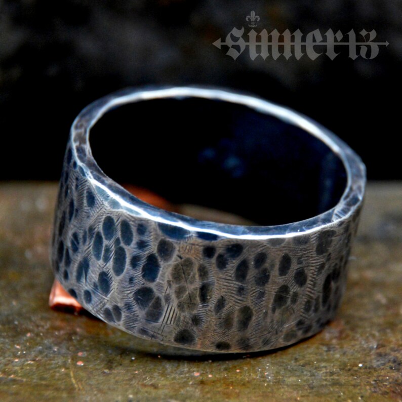 May include: A wide silver band ring with a hammered texture. The ring has a dark, almost black, oxidised finish.