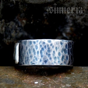 May include: A silver ring with a hammered texture. The ring is wide and has a flat top.