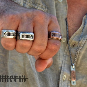 May include: Three silver and copper rings with the word "SUMMER" engraved on them. The rings are worn on the fingers of a person's hand. The person is also wearing a silver chain necklace with a copper pendant that also has the word "SUMMER" engraved on it.