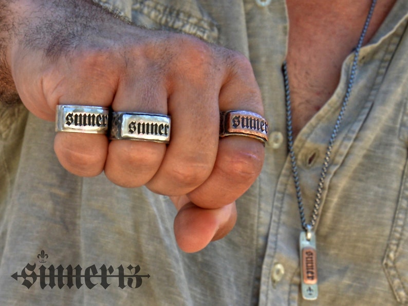 May include: Three silver and copper rings with the word "SUMMER" engraved on them. The rings are worn on the fingers of a person's hand. The person is also wearing a silver chain necklace with a copper pendant that also has the word "SUMMER" engraved on it.