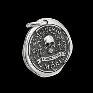 Könnte beinhalten: Ein runder silberner Anhänger mit einem zentralen Totenkopfmotiv, umgeben von kunstvollen Verzierungen und zwei Kreuzen. Der Text MEMENTO MORI ist am Rand eingeprägt, mit einem Banner in der Mitte, das die Worte CARPE DIEM zeigt.