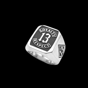 Puede incluir: Un anillo de metal plateado con una cara rectangular y un fondo negro hundido. La cara muestra el número 13 en el centro, rodeado por el texto LOYALTY arriba y RESPECT abajo. En el lateral hay una pequeña forma de diamante con el número 13.