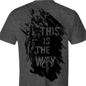 Puede incluir: Camiseta gris oscuro con un dise&ntilde;o gr&aacute;fico negro. El dise&ntilde;o presenta una figura con casco, el texto "THIS IS THE WAY" y una calavera estilizada. El dise&ntilde;o tiene un efecto de pincelada.